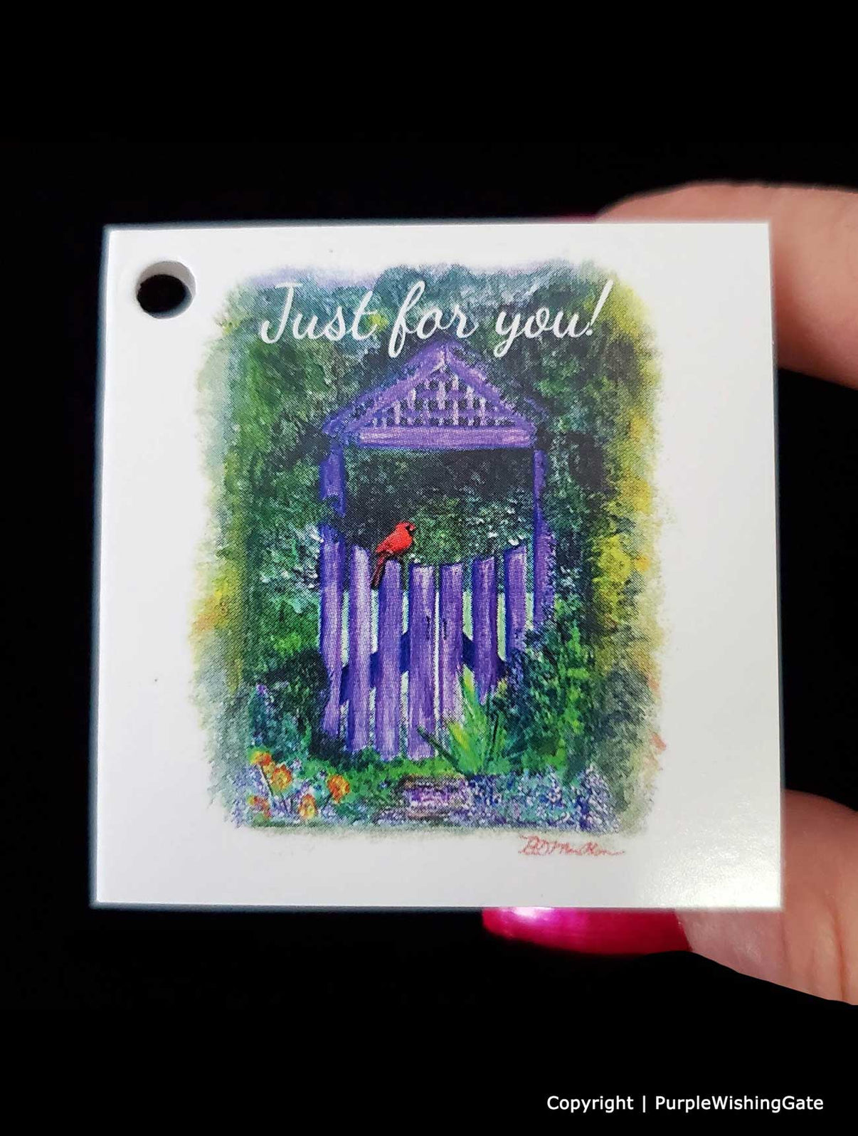Gift Tag with a purple gate and red bird illustration, held by a hand against a black background.