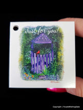 Gift Tag with a purple gate and red bird illustration, held by a hand against a black background.